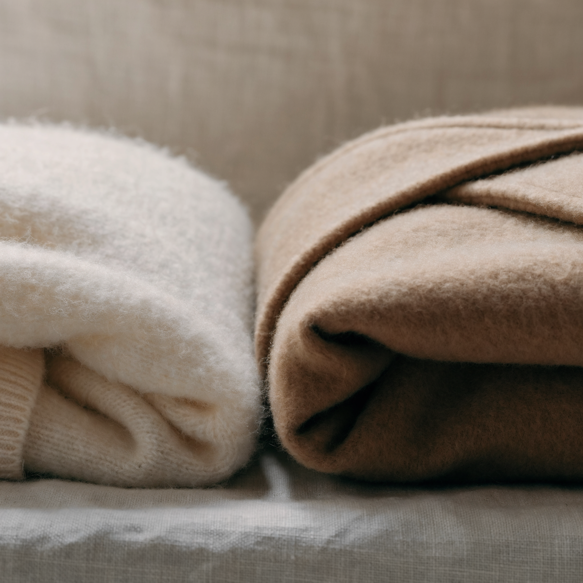 Folded cream knitwear and a camel wool coat on a linen surface