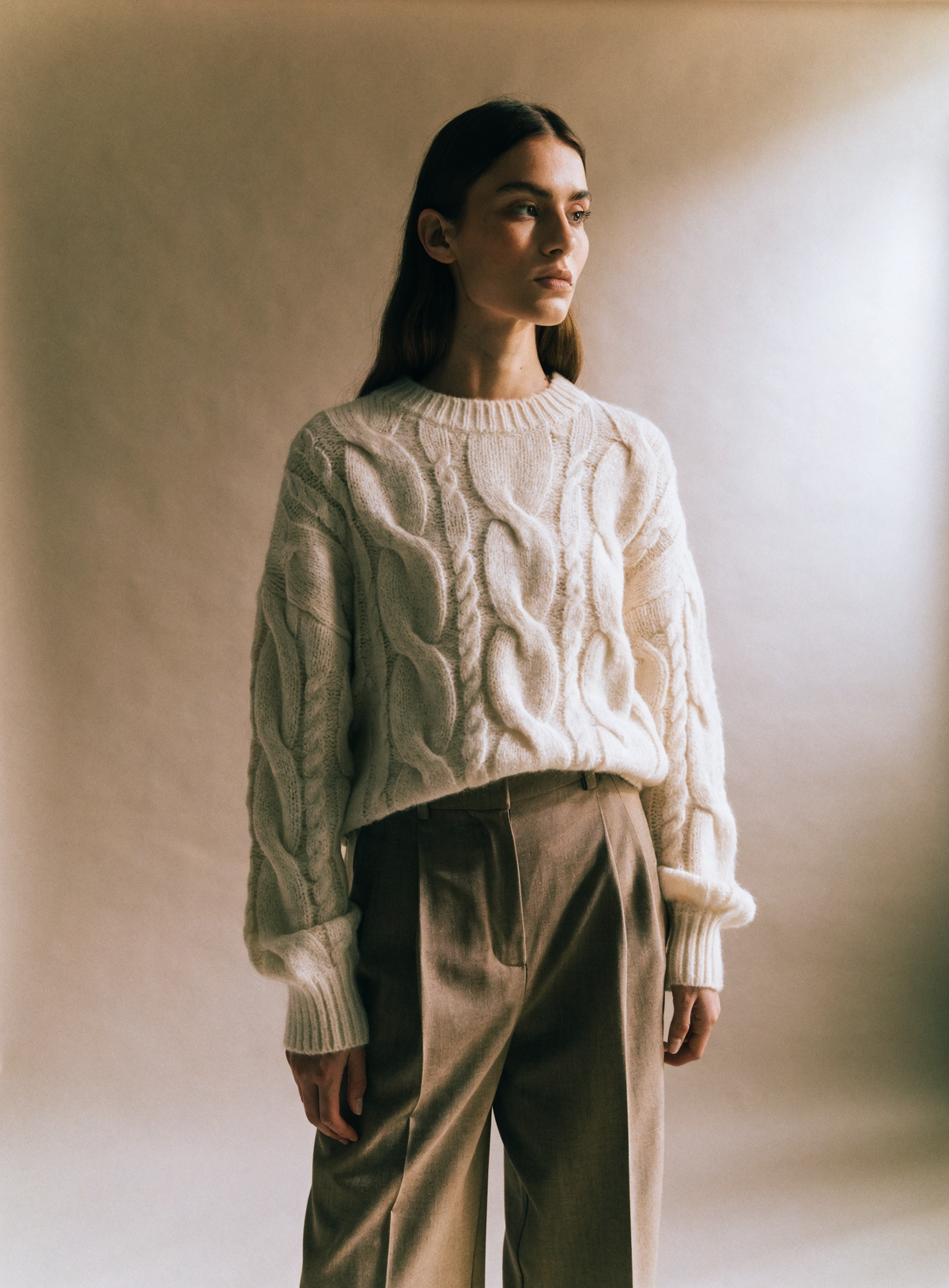 Model in a cream cable-knit sweater and tailored trousers against a warm neutral studio backdrop